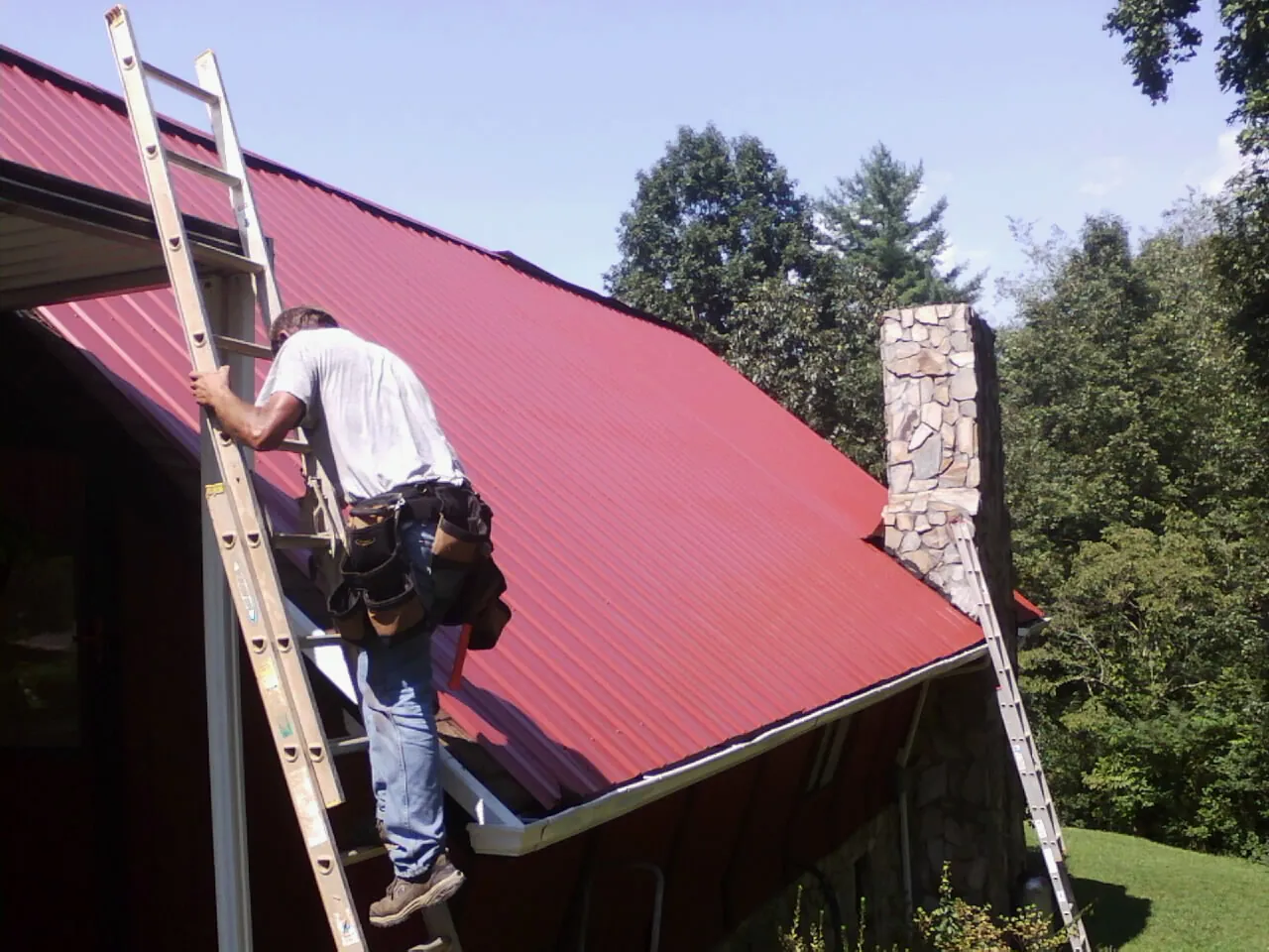 Roofer on a ladder beside a red metal roof and stone chimney — Soffit & Fascia Repair by Roofing Brothers in Duncan
